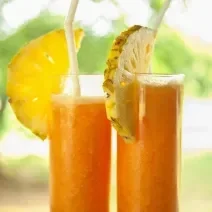 Fotografia de dois copos de vidro grandes com suco de laranja, abacaxi e mel. Em cada copo, tem metade de uma fatia de abacaxi e tem também um canudo. Ao fundo tem uma paisagem com de natureza com árvores.