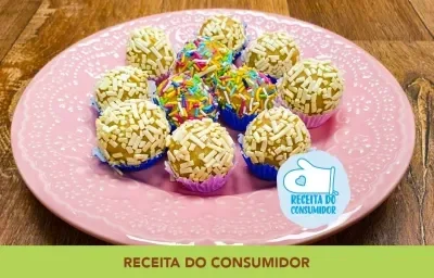 Imagem aproximada de um prato em tom de rosa, sobre uma bancada de madeira. Nele há onze brigadeiros em tons brancos, beges e com granulados brancos e coloridos, todos em forminhas coloridas