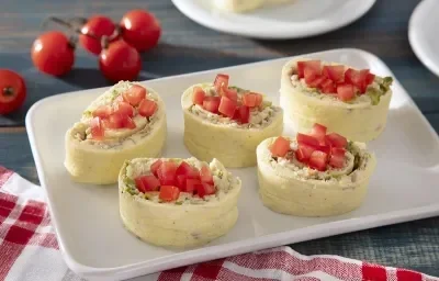 Foto em tons claros da receita de rolinho de ovos servida em 5 porções sobre uma base de porcelana branca. Ao fundo há um ramo de tomates-cereja e ao lado um paninho quadriculado vermelho e branco