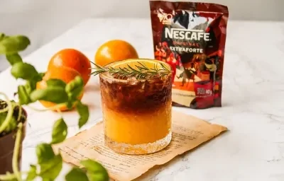 Fotografia em tons de laranja em uma bancada de mármore com um copo de vidro ao centro e o suco de laranja com café dentro dele, decorado com um ramo de alecrim. Ao fundo, laranja e embalagem de café. Fotografia em tons de laranja em uma bancada de mármore com um copo de vidro ao centro e o suco de laranja com café dentro dele, decorado com um ramo de alecrim. Ao fundo, laranja e embalagem de café.
