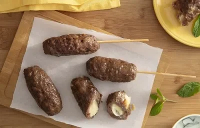 Foto da receita de Kafta Recheada com queijo. Observa-se 4 kaftas: duas no espeto, uma na tábua e outra cortada ao meio, expondo o recheio cremoso de queijo. Estão sobre uma tábua de madeira e há hortelãs decorando a foto. Foto da receita de Kafta Recheada com queijo. Observa-se 4 kaftas: duas no espeto, uma na tábua e outra cortada ao meio, expondo o recheio cremoso de queijo. Estão sobre uma tábua de madeira e há hortelãs decorando a foto.