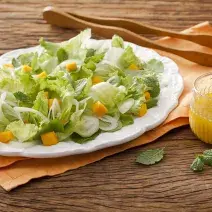 Fotografia em tons de marrom em uma bancada de madeira com um pano laranja e um prato branco oval com as hortaliças. Ao lado, um pegador de salada de madeira e um copo de vidro com suco de laranja.