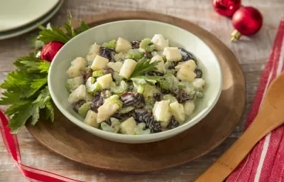 Foto da receita de salpicão de ameixa e iogurte servida em um bowl verde claro sobre uma tábua de madeira circular em cima de uma mesa decorada no tema de natal com uma colher de madeira ao lado