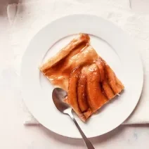 Fotografia vista de cima de uma torta de banana com bananas caramelizadas por cima em um prato de sobremesa branco, e uma colher ao lado. A sobremesa está em cima de um pano branco, que está sobre uma mesa branca.