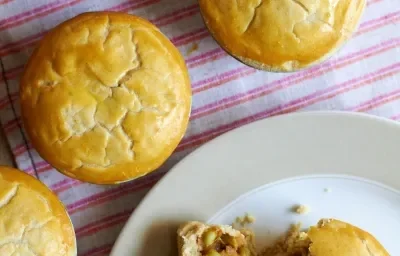 Fotografia em tons de laranja em uma mesa de madeira com um pano listrado em vermelho e empadinhas de palmito espalhadas no pano. Ao lado, um prato raso branco com uma empadinha cortada ao meio.