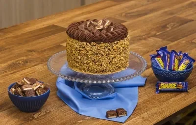 Foto aproximada da receita de Bolo Charge, decorado com amendoins e chocolate, sobre uma bancada de madeira, com alguns bombons Charge ao redor Foto aproximada da receita de Bolo Charge, decorado com amendoins e chocolate, sobre uma bancada de madeira, com alguns bombons Charge ao redor