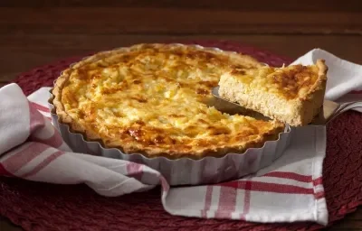 Fotografia em tons de marrom e roxo de uma bancada de madeira vista de cima, ao centro uma forma de alumínio com uma torta dentro, acima uma espátula com uma fatia e abaixo da forma um pano listrado nas cores vermelho e branco.