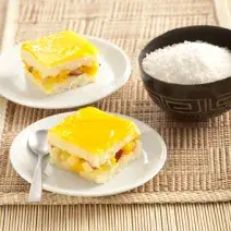 Fotografia em tons de amarelo em uma bancada de madeira com uma toalha de palha, dois pratos brancos redondos com um pedaço de pavê de tapioca em cada um. Ao lado, um potinho preto com coco ralado.