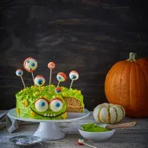 Imagem escurecida de um bolo temático de monstro em tons verde e marrom com decorações de dia das bruxas, sobre uma bancada cinza com algumas abóboras ao fundo