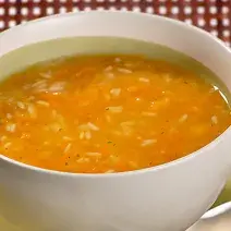 Fotografia em tons de vermelho e laranja em uma mesa com uma toalha vermelha, um prato redondo fundo com a sopa de abóbora dentro dele.