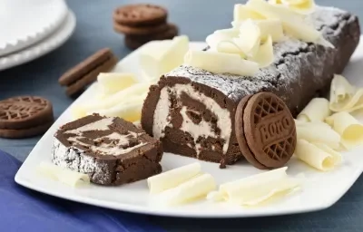 Fotografia em tons de azul em uma bancada de madeira azul, um pano azul escuro, um prato quadrado branco raso com o rocambole de chocolate com recheio de Leite Moça e raspas de chocolate branco em cima do prato. Ao lado, Biscoitos Negresco espalhados.