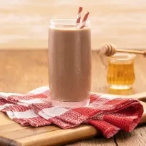 Foto da receita de milkshake proteico com abobrinha e cacau servida em um copo alto e largo com dois canudos listrados, embaixo um pano quadriculado vermelho e branco sobre uma tábua de madeira. Ao fundo um copo do vidro com mel