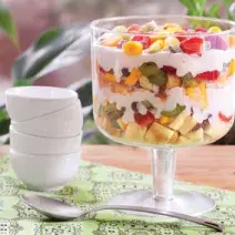 em uma bancada contém uma taça de vidro com pedaços de frutas e creme por cima e ao lado uma colher e potinhos brancos empilhados.