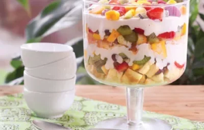 em uma bancada contém uma taça de vidro com pedaços de frutas e creme por cima e ao lado uma colher e potinhos brancos empilhados.