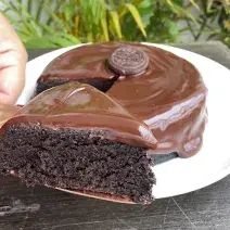 Foto aproximada de um bolo de chocolate com a fatia na frente sobre uma faca. O bolo está decorado com uma calda de chocolate e com um biscoito Negresco no centro.