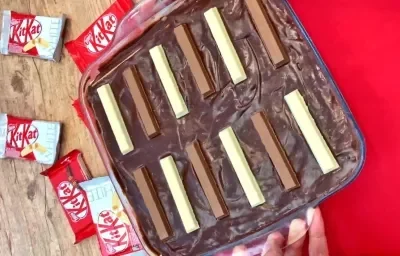 Foto de um pavê de KitKat ao leite e branco, numa travessa de vidro, sobre um tecido vermelho com alguns pacotes de KitKat ao fundo.