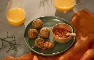 Foto em tons de laranja e verde escuro da receita de bolinho de arroz e aveia servido em 4 porções com uma cumbuca de geleia de pimenta ao lado, tudo sobre um prato de cerâmica verde. Ao lado há um paninho laranja e dois copos de suco de laranja