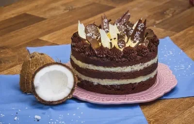 Foto da receita de Bolo Naked de Prestígio, decorado com pedaços de chocolate, sobre um prato rosa, numa bancada de madeira com tecidos azuis e dois pedaços de coco fresco aberto Foto da receita de Bolo Naked de Prestígio, decorado com pedaços de chocolate, sobre um prato rosa, numa bancada de madeira com tecidos azuis e dois pedaços de coco fresco aberto