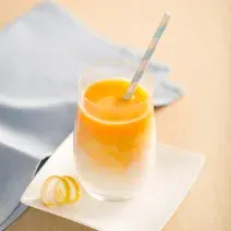 Fotografia em tons de laranja e azul em uma bancada de madeira clara, um pano azul, um prato pequeno branco quadrado com um copo de vidro em cima dele e o smoothie de laranja, manga e cenoura dentro dele.