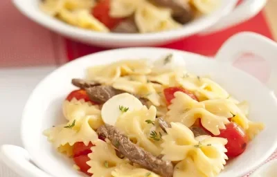 Fotografia em tons de vermelho em uma bancada de madeira, um jogo americano vermelho e branco, um prato branco redondo com o macarrão com tirinhas de carne e tomate.