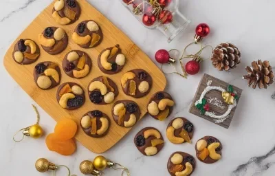 Foto vista de cima da receita de moedas de chocolate com frutas secas e nuts, sobre uma tábua de madeira, há algumas sobre a bancada de mármore onde também há pinhas, bolas de natal, uma plaquinha natalina e alguns damascos soltos