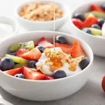 Fotografia em tons de branco, com três refratários brancos fundos no canto e no centro outro refratário com diversas frutas, iogurte e uma calda de mel caindo. No canto inferior direito, dois morangos inteiros.