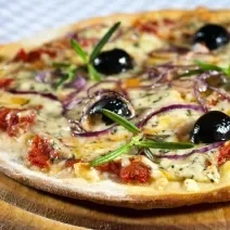 Fotografia de uma pizza de massa fina com mozarela, gorgonzola, requeijão, tomate seco, orégano, cebola roxa e azeitonas pretas. A pizza está sobre um apoio de madeira.