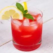 Fotografia em tons de vermelho em uma bancada de madeira de cor branca. Ao centro, um copo contendo a bebida pink lemonade.
