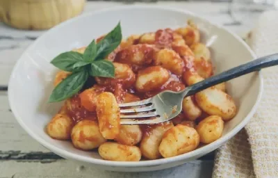 Foto da receita de Nhocão da Gisele. Observa-se um prato fundo branco com os nhoques dentro, um ramo de manjericão decorando e um garfo espetando um nhoque. Foto da receita de Nhocão da Gisele. Observa-se um prato fundo branco com os nhoques dentro, um ramo de manjericão decorando e um garfo espetando um nhoque.
