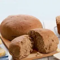 Fotografia em tons de marrom em uma bancada de madeira com uma tábua de madeira clara e o pão preto redondo grane apoiado nela, e um outro pedaço de pão já cortado.