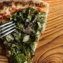 Fotografia em tons de verde em uma bancada de madeira de cor marrom. Ao centro, uma fatia de pizza de escarola com um garfo ao lado.