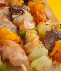 Foto com um fundo amarelo e variações de carnes como filé mignon, frango, lombo suíno, vegetais colocados em um espeto