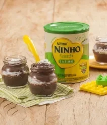 Receitinhas com Ninho Sobremesa de prestígio em potinhos sobre bancada clara com pano verde xadrez, brinquedos e lata de Ninho Fases ao lado.