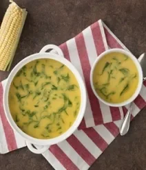 Fotografia em tons de vermelho e amarelo, com um pano listrado vermelho e em cima dele uma panela branca com sopa de milho
