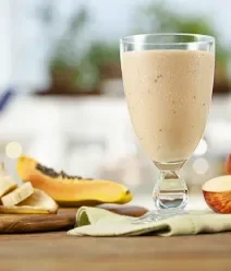 Vitamina cremosa em taça, cercada por banana, mamão e maçã sobre mesa de madeira.