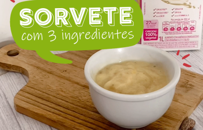 Foto da receita de sorvete de banana servida em um bowl de porcelana branco sobre uma tábua de madeira com amêndoas ao lado direito e bananas em rodelas ao lado esquerdo. Ao fundo, há uma caixa da bebida vegetal de amêndoas nature's heart