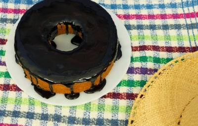 Um bolo de cenoura inteiro com cobertura de chocolate, no formato redondo com furo no meio, está em um prato branco sobre uma toalha colorida xadrez. Um chapéu de palha aparece parcialmente no canto da imagem.