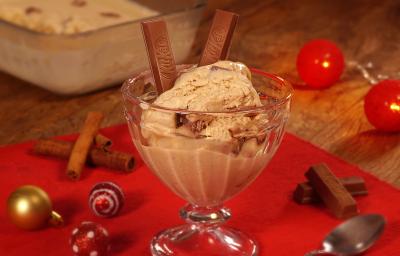 Fotografia em tons de marrom e vermelho de uma bancada de madeira com paninho vermelho, sobre ele uma taça com o sorvete. Ao lado bolinhas natalinas, uma colher, paus de canela e kitkat. Ao fundo um recipiente com o sorvete.