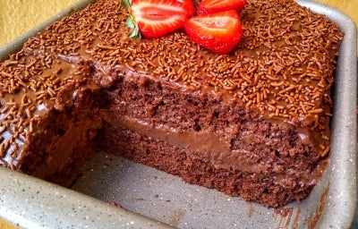 Fotografia em tons claros de uma forma retangular, antiaderente, com o bolo de chocolate, recheado e coberto, finalizado com granulado e pedaços de morango no centro. A foto mostra cerca de dois pedaços do bolo faltando, o que foca na camada do bolo. Fotografia em tons claros de uma forma retangular, antiaderente, com o bolo de chocolate, recheado e coberto, finalizado com granulado e pedaços de morango no centro. A foto mostra cerca de dois pedaços do bolo faltando, o que foca na camada do bolo.