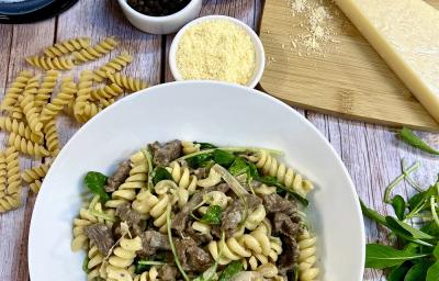 Foto da receita de fusilli com filé mignon e rúcula servida em um prato de porcelana branco sobre uma mesa de madeira. Como decoração, há folhas de rúcula a direita e macarrão a esquerda, além de recipientes com queijo ralado e ingredientes da receita