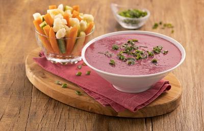 Foto da receita de pasta de beterraba com legumes servida em um bowl de porcelana branca sobre um pano roxo com um recipiente de vidro com legumes variados em fatias ao lado, tudo sobre uma tábua de madeira. Ao fundo, um recipiente de vidro com cebolinha