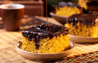 Fotografia em tons de marrom com uma prato pequeno ao centro. Em cima do prato existe uma fatia de um bolo de cenoura de cor alaranjada e com cobertura de chocolate. Ao fundo existe mais fatias desse mesmo bolo. Fotografia em tons de marrom com uma prato pequeno ao centro. Em cima do prato existe uma fatia de um bolo de cenoura de cor alaranjada e com cobertura de chocolate. Ao fundo existe mais fatias desse mesmo bolo.