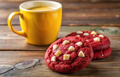 Fotografia de três cookies de red velvet com gotas de chocolate branco juntos, sobre uma mesa de madeira escura, ao lado de uma xícara grande amarela. Fotografia de três cookies de red velvet com gotas de chocolate branco juntos, sobre uma mesa de madeira escura, ao lado de uma xícara grande amarela.