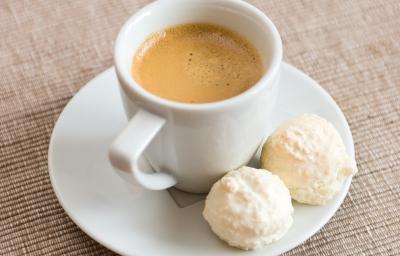 fotografia em tons de branco, cinza e marrom de uma bancada cinza. Contém um pratinho redondo branco com uma xicara de café branca com café dentro, ao lado dois bolinhos redondos.