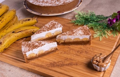 Em cima de uma mesa, fatias de banoffee com massa de biscoitos crocante, doce de leite, bananas em rodelas, decorado com chantilly cremoso, polvilhado canela em pó.