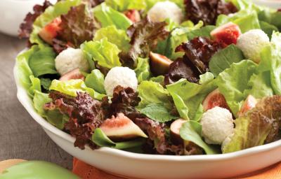 Uma bancada de madeira cinza vista de cima contém um pano laranja, um recipiente redondo e branco com salada dentro pedaços de figo e bolinhas de ricota. À frente duas colheres verdes para mexer a salada.