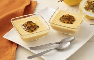Fotografia em tons de laranja em uma bancada de madeira clara com um pano laranja, ao centro um prato branco quadrado e duas taças de vidro com o flan de maracujá dentro, enfeitados com sementes de maracujá. Ao fundo, um maracujá cortado ao meio. Fotografia em tons de laranja em uma bancada de madeira clara com um pano laranja, ao centro um prato branco quadrado e duas taças de vidro com o flan de maracujá dentro, enfeitados com sementes de maracujá. Ao fundo, um maracujá cortado ao meio.