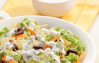 Fotografia em tons de tons de branco, amarelo e verde, saladeira com folhas verdes, azeitona, cenoura e molho de iogurte sobre guardanapo amarelo, ao lado potinho com molho de iogurte e folhas de hortelã, tudo sobre bancada em tom pastel. Fotografia em tons de tons de branco, amarelo e verde, saladeira com folhas verdes, azeitona, cenoura e molho de iogurte sobre guardanapo amarelo, ao lado potinho com molho de iogurte e folhas de hortelã, tudo sobre bancada em tom pastel.