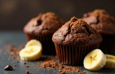 Três bolinhos de chocolate estão em destaque sobre uma superfície escura, decorada com fatias de banana e farelos de cacau ao redor. A textura dos bolinhos é visivelmente macia e úmida, com uma aparência caseira e aconchegante. Três bolinhos de chocolate estão em destaque sobre uma superfície escura, decorada com fatias de banana e farelos de cacau ao redor. A textura dos bolinhos é visivelmente macia e úmida, com uma aparência caseira e aconchegante.