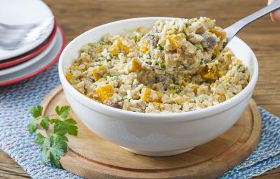 Fotografia em tons de branco, amarelo e marrom, com bowl branco ao centro sobre tábua de cozinha, ramo de salsa e um guardanapo azul, com arroz cremoso com legumes e carne picada, ao fundo pratos com garfos por cima, tudo sobre bancada de madeira marrom. Fotografia em tons de branco, amarelo e marrom, com bowl branco ao centro sobre tábua de cozinha, ramo de salsa e um guardanapo azul, com arroz cremoso com legumes e carne picada, ao fundo pratos com garfos por cima, tudo sobre bancada de madeira marrom.
